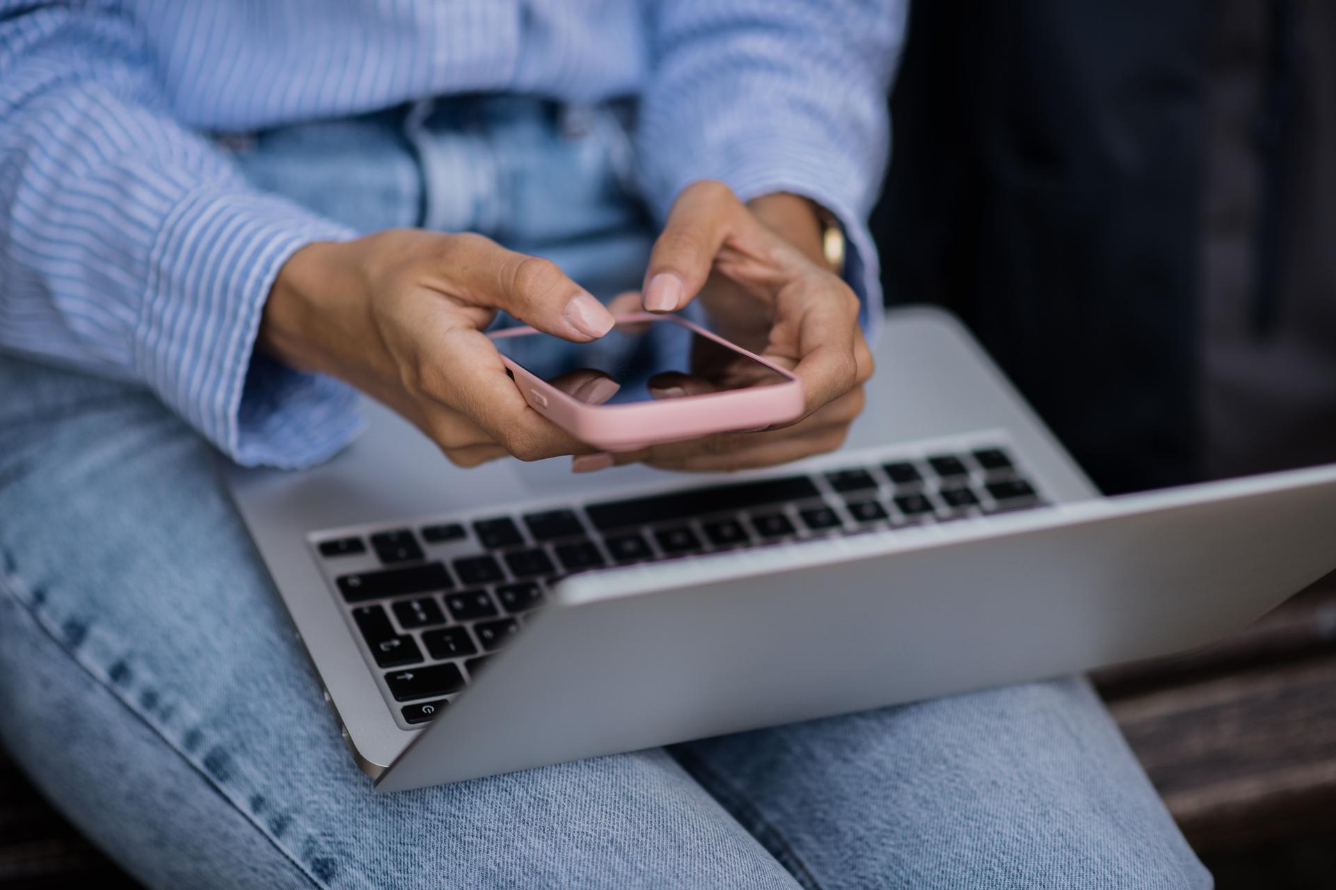 Close-up of young woman typing on phone with laptop on lap