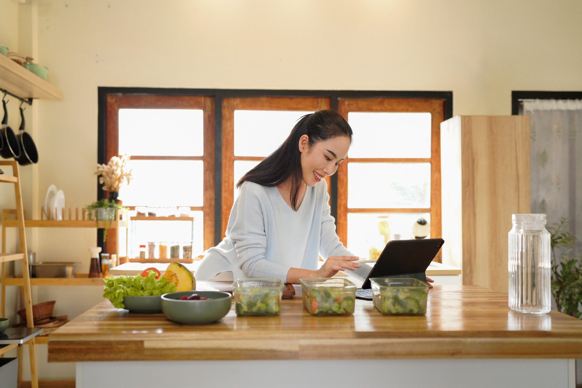 Woman organizing kitchen containers while using a tablet for meal planning on counter.